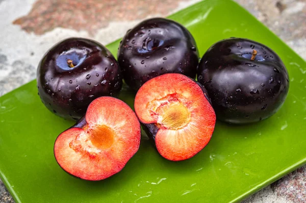 Tres frutos de ciruela morados oscuros y uno cortado en un plato verde — Foto de Stock