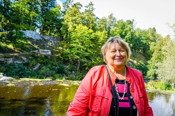 Mogen kvinna i en röd jacka som vilar vid älven. — Stockfoto