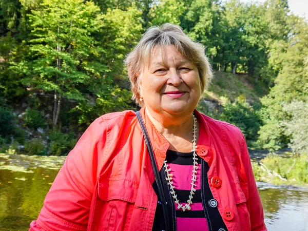 Volwassen vrouw in rode jas rust in het park aan de rivier. — Stockfoto