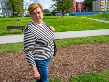 Çizgili tişörtlü olgun bir kadın parkta yürüyor.