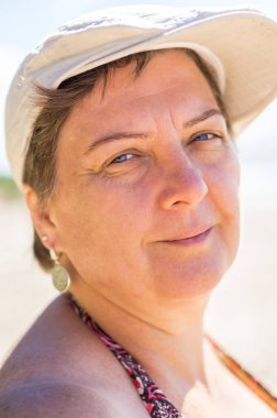 Olgun, tombul bir kadın, mayolu ve kepli deniz kenarında dinleniyor.