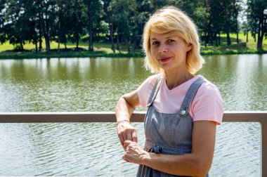 Orta yaşlı bir kadın şehir parkında bir gölün kıyısında yürüyor.