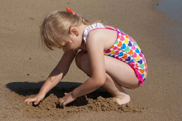 Kleines Mädchen im bunten Bikini, das am Strand mit Sand spielt Stockbild