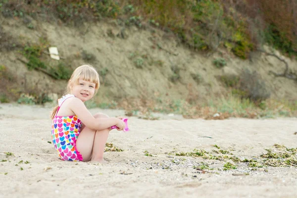 Niño juega sentado en la arena en la playa en un día soleado de verano. Niña feliz . — Foto de Stock