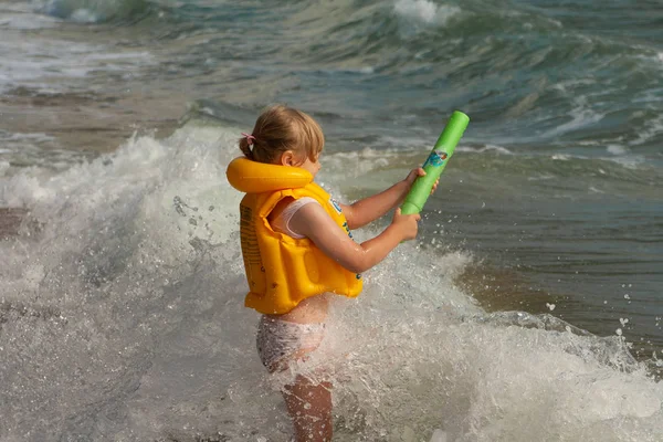 Chica joven con chaleco salvavidas amarillo. En la orilla con las olas. Pistola de agua . — Foto de Stock