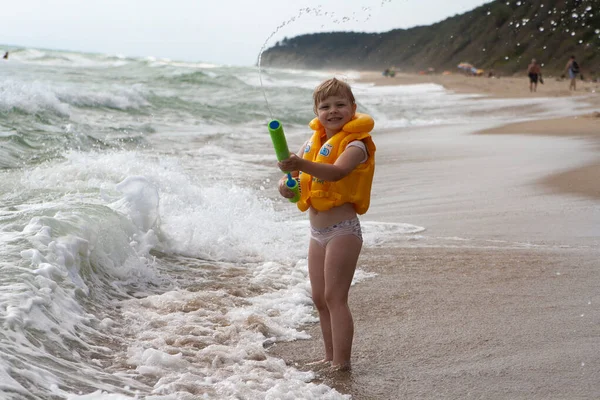 Ein kleines Mädchen in gelber Schwimmweste am Strand spielt mit einem Wasserwerfer. — Stockfoto