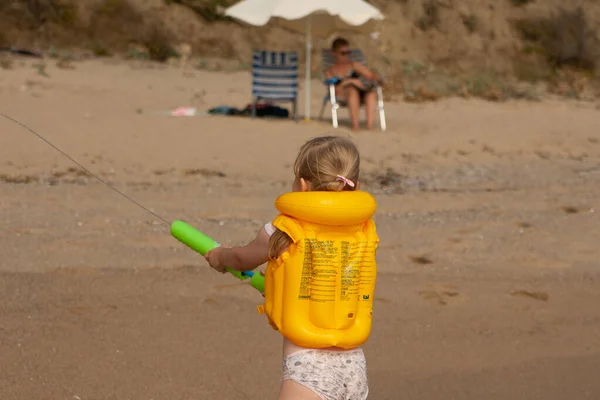 Una niña con un chaleco salvavidas amarillo en la playa juega con un cañón de agua . — Foto de Stock