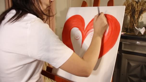 年轻的女艺术家画一个红色的心脏与丙烯酸油漆在一个白色的画布上的画架在她的艺术工作室 — 图库视频影像