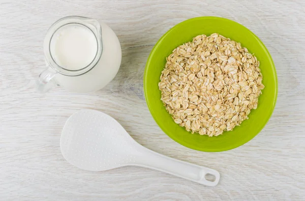 Dry oat flakes in bowl, jug of milk and plastic spoon on wooden table. Top view