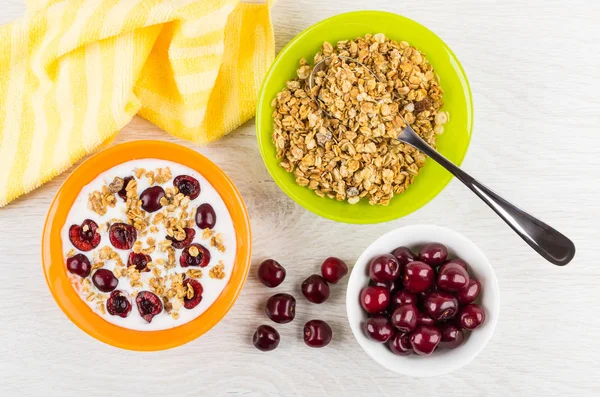 Muesli with cherries, yogurt in bowl, spoon in bowl with muesli, cherry, napkin, spoon on wooden table. Top view