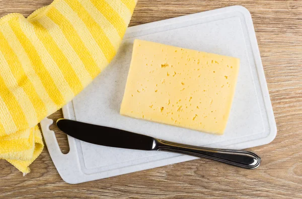 Piece of cheese, table knife on plastic cutting board, yellow napkin on wooden table. Top view