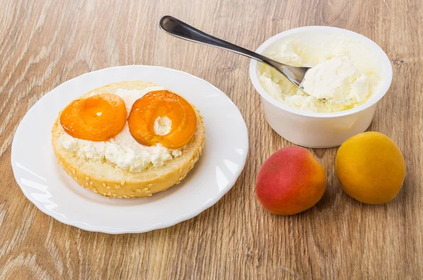 Sandwich with cream cheese, apricot in white plate, plastic jar with cheese, spoon, apricots on wooden table