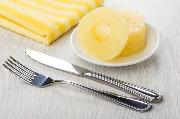Rings of canned pineapple in white saucer, napkin, knife and fork on wooden table