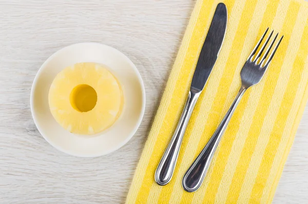 Rings of canned pineapple in saucer, fork, knife on yellow napkin on wooden table. Top view