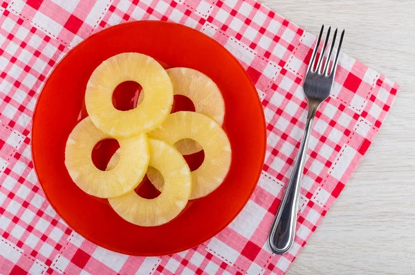 Rings of canned pineapple in red plate, fork on checkered napkin on wooden table. Top view