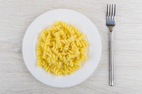 Boiled pasta fusilli in white plate, fork on wooden table. Top view