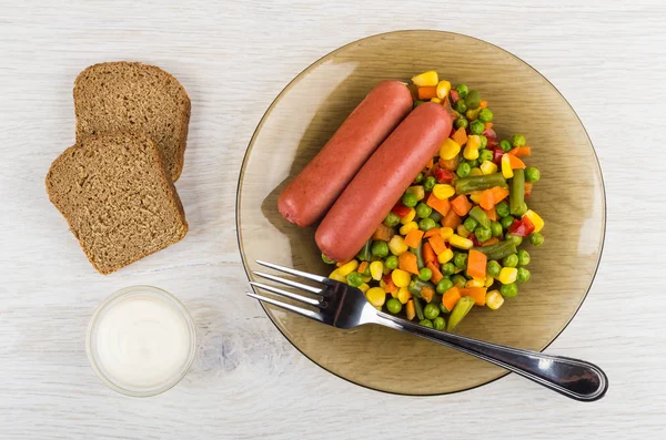 Plate with boiled sausages, vegetable mix, pieces of bread, bowl with mayonnaise, fork on wooden table. Top view