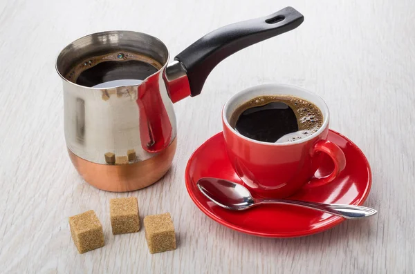 Boiled coffee in cezve, coffee in red cup, spoon on saucer, sugar cubes on wooden table