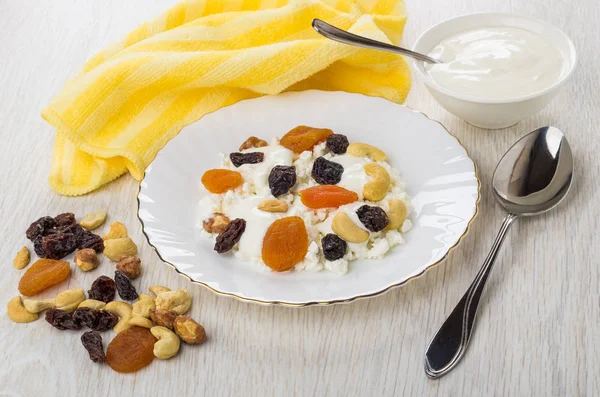 Cottage cheese, yogurt, dried fruits, nuts in plate, spoon, heap of fruits, nuts, napkin on wooden table