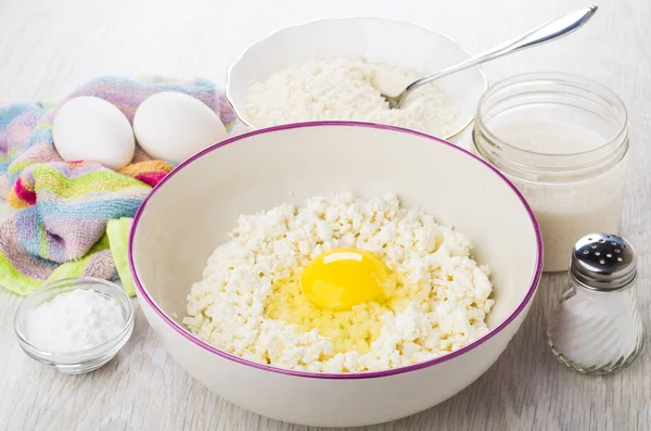 Cottage cheese with egg in bowl, eggs on napkin, soda, sugar, salt, flour in bowl on wooden table