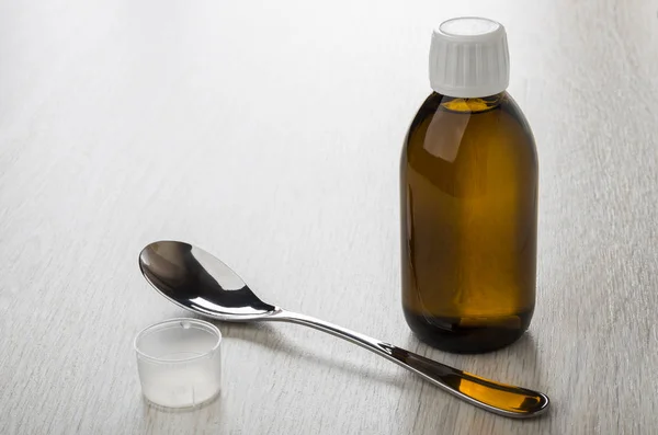 Cough syrup in brown transparent bottle, spoon and measuring cap on wooden table