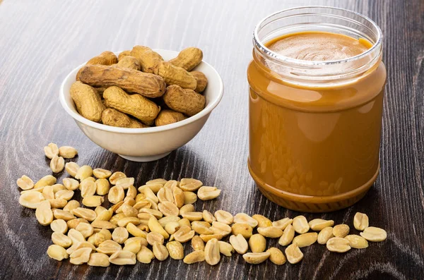 Peanut paste in open plastic jar, bowl with nuts in shell, fried peanuts on wooden table