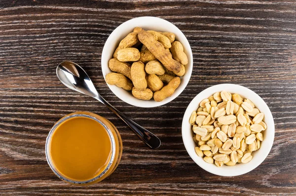 Peanut paste in open jar, spoon, bowls with fried nuts in shell and peeled peanuts on wooden table. Top view