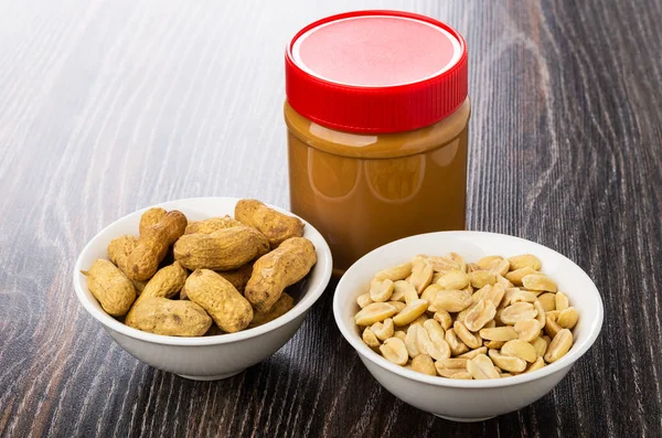 Peanut paste in plastic jar, bowls with fried nuts in shell and peeled peanuts on wooden table