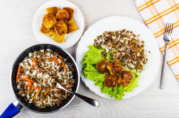 Cooked mushroom, lentils, rice, spoon in frying pan, fried nuggets in plate, dish with vegetable mix and nuggets, fork, napkin on wooden table. Top view