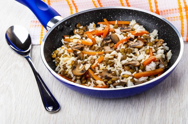 Cooked mix from mushroom, lentils, rice in frying pan, napkin, spoon on wooden table