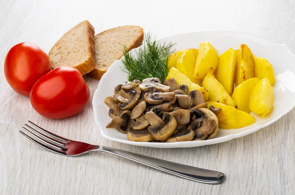 Pieces of fried potato, mushroom, dill in white plate, red tomatoes, slices of bread, fork on wooden table