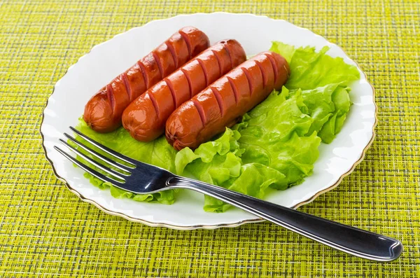 Grilled sausages, lettuce, fork in white plate on green mat