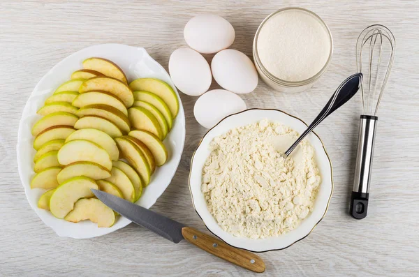 Slices of apples in plate, flour in bowl, eggs, sugar in jar, whisk, kitchen knife on wooden table. Top view