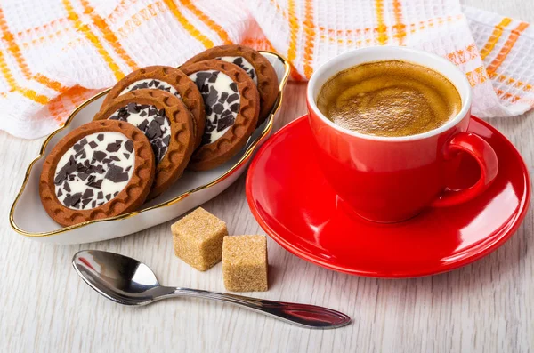 Round cookies with cream and chocolate in white plate, coffee espresso in red cup on saucer, sugar cubes, spoon, napkin on wooden table