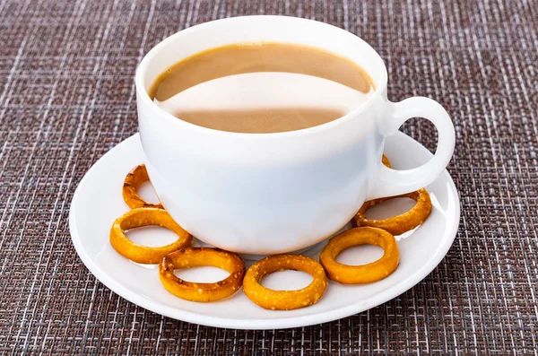 Cup of cocoa with milk, salted bread rings around cup on saucer on dark mat