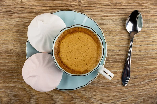 Two marshmallows, coffee espresso in cup on saucer, spoon on wooden table. Top view