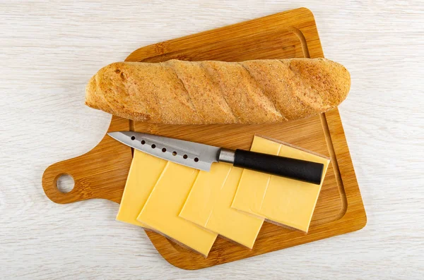 Loaf of bread, packages of melted cheese, kitchen knife on bamboo cutting board on wooden table. Top view