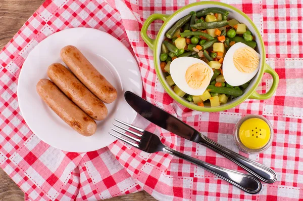 Cooked vegetable mix, halves of boiled egg in bowl, pepper shaker, fried sausages in plate, fork and knife on checkered napkin on wooden table. Top view
