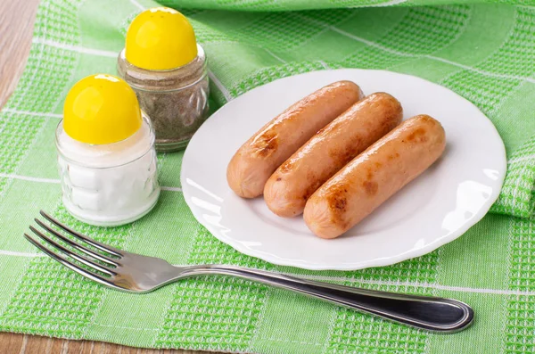 Salt shaker, pepper shaker, fried sausages in white plate, fork on green napkin on wooden table