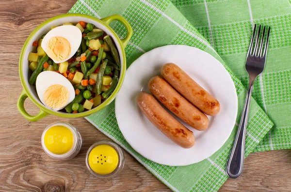 Cooked vegetable mix, halves of boiled egg in bowl, knife, salt shaker, pepper shaker, fried sausages in plate, fork on green napkin on wooden table. Top view