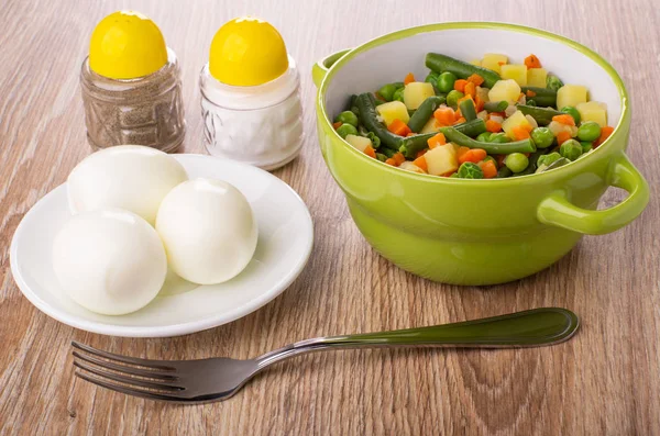 Cooked vegetable mix with green bean, green peas, potato, onion, carrot in bowl, salt shaker, pepper shaker, boiled eggs in saucer and fork on wooden table