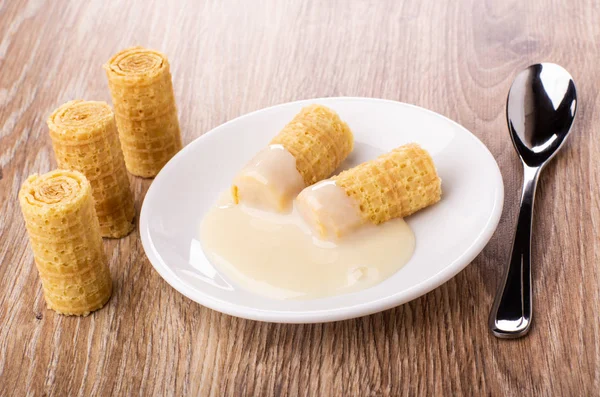 Wafer rolls with condensed milk in saucer, rolls and teaspoon on wooden table