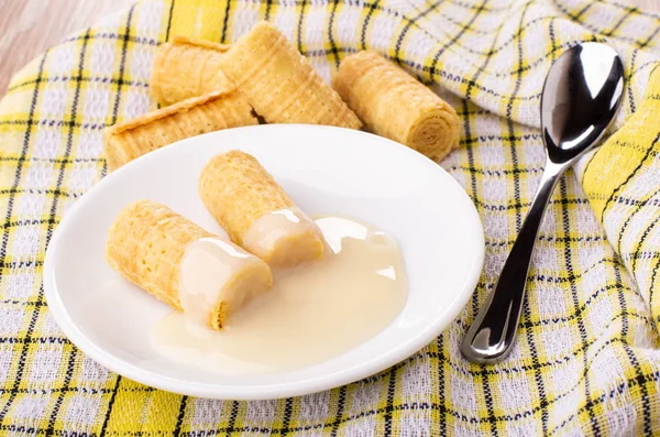 Wafer rolls with condensed milk in saucer, wafer rolls on checkered napkin on wooden table