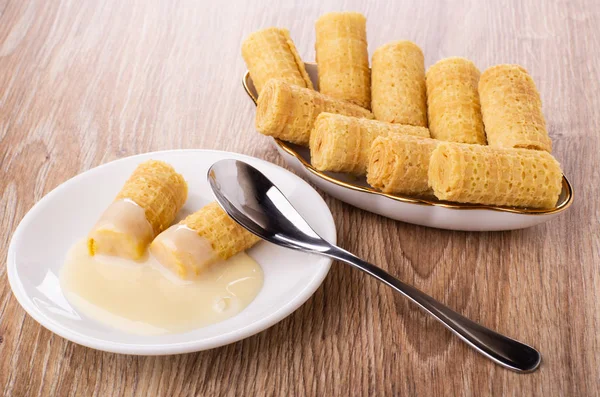 Wafer rolls with condensed milk, spoon in saucer, wafer rolls in plate on wooden table