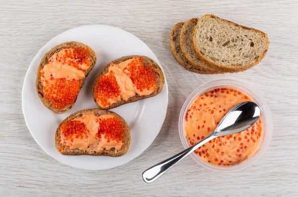 Sandwiches with caviar in white plate, spoon, imitation of red caviar with cream in plastic jar, pieces of bread on wooden table. Top view