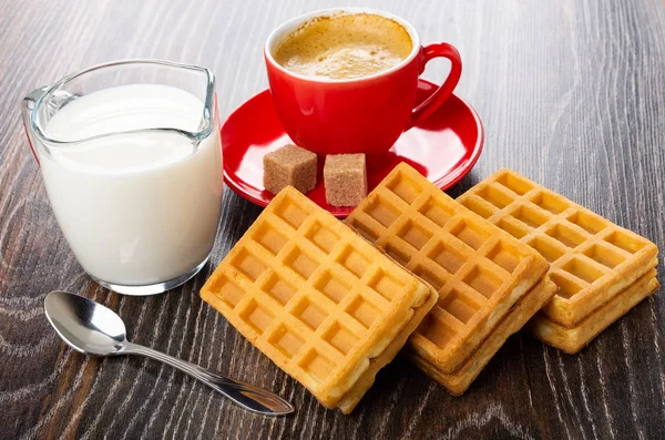Pitcher with milk, soft waffles with filling, red cup with coffee espresso, sugar cubes on saucer, spoon on dark wooden table