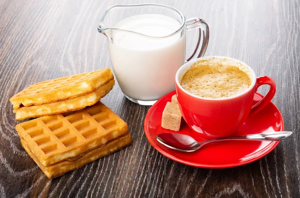 Soft waffles with filling, pitcher with milk, red cup with coffee espresso, sugar cubes, spoon on saucer on dark wooden table