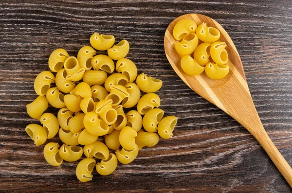 Heap uncooked pasta lumache, pasta in bamboo spoon on wooden table. Top view