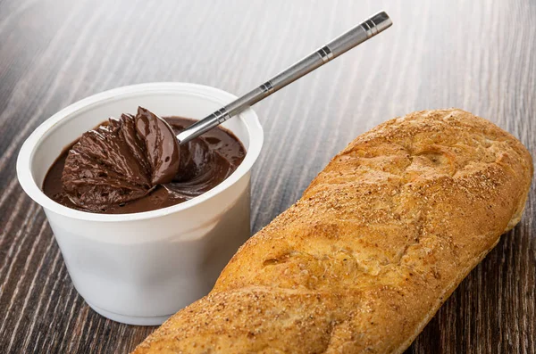 Spoon with chocolate melted cheese in plastic jar, loaf of bread