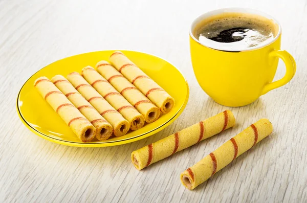Striped wafer rolls in yellow glass saucer, cup of black coffee on light wooden table